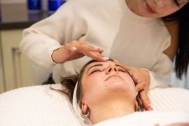 Woman receiving a facial treatment. Another person's hands gently touch her forehead. White sweater, bright setting.