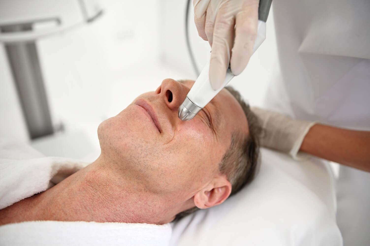 Man receiving a facial treatment in a bright room. A gloved hand holds a device near his face.