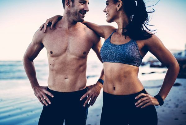 Fit couple at the beach: man shirtless with hand on hip, woman in sports bra, both smiling.