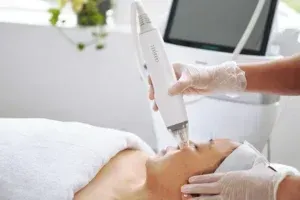 Person receiving facial laser treatment in a spa setting. A device is held close to the face by gloved hands.