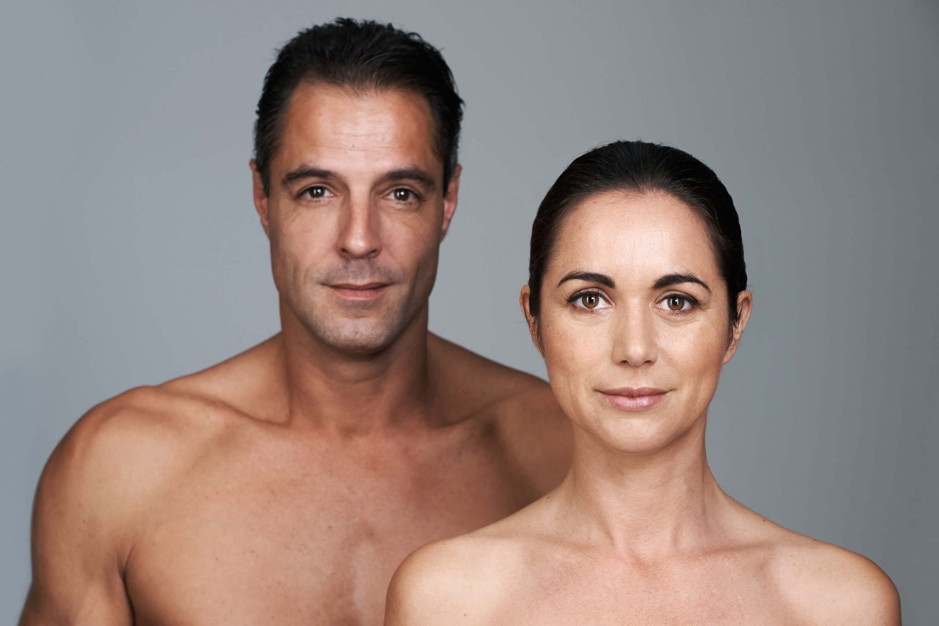 Man and woman with bare shoulders, smiling in front of a gray backdrop.