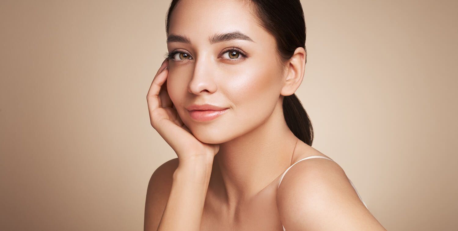 Woman with fair skin touches cheek, looking toward the viewer against a neutral background.
