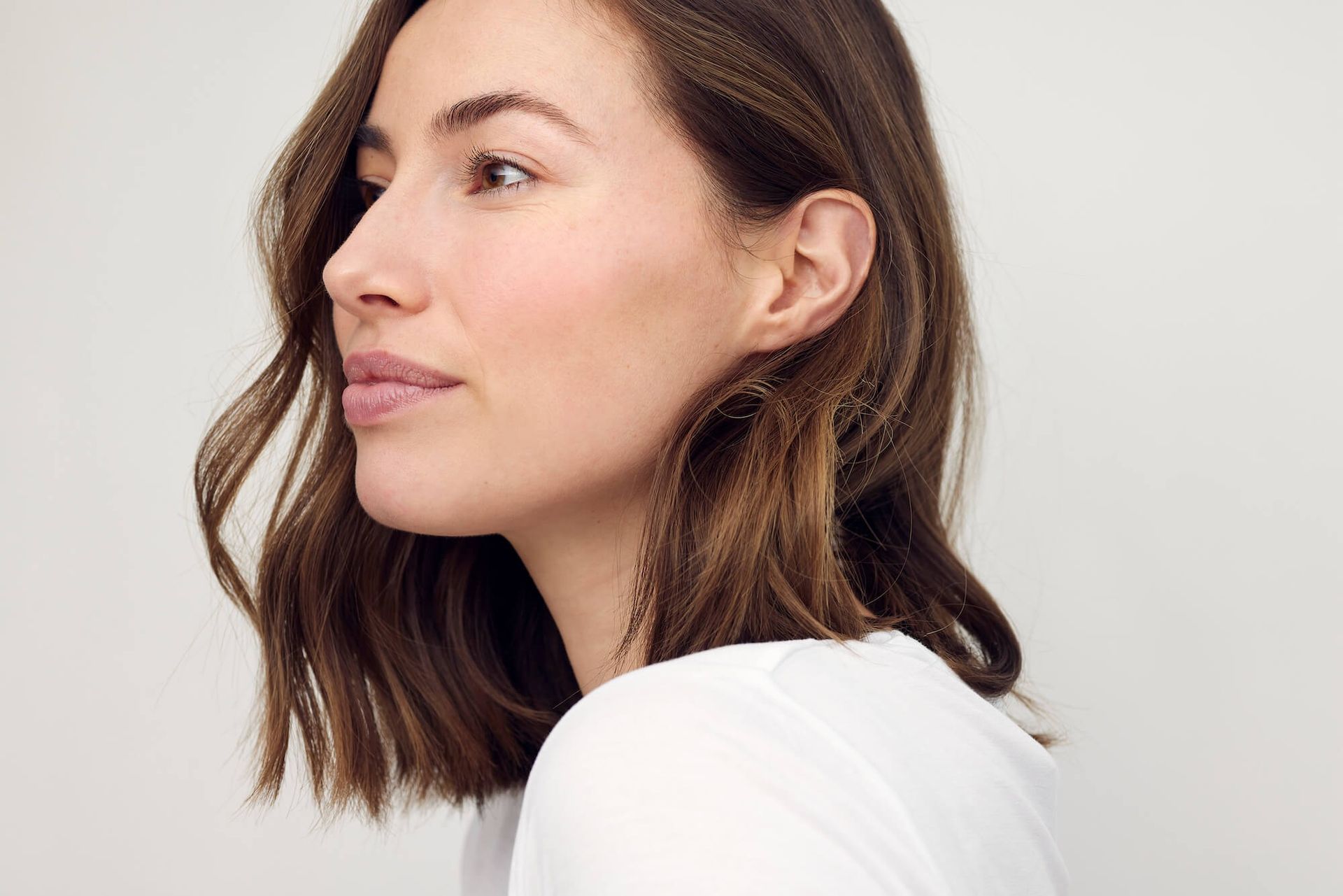 Woman with brown, wavy hair looking to the side, wearing a white shirt against a light background.