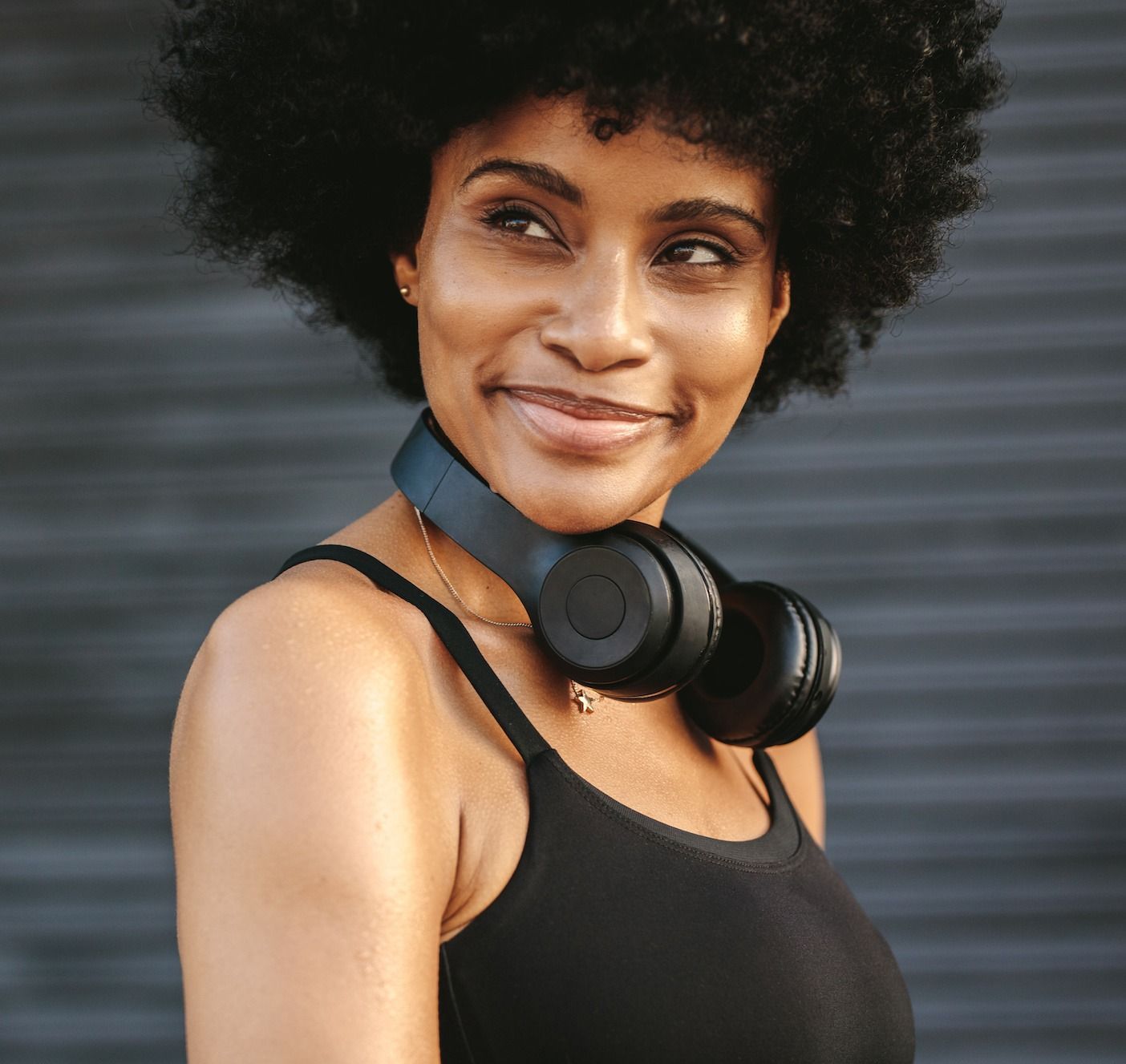 Woman with headphones around neck, looking to the side, smiling. Black tank top, dark background.