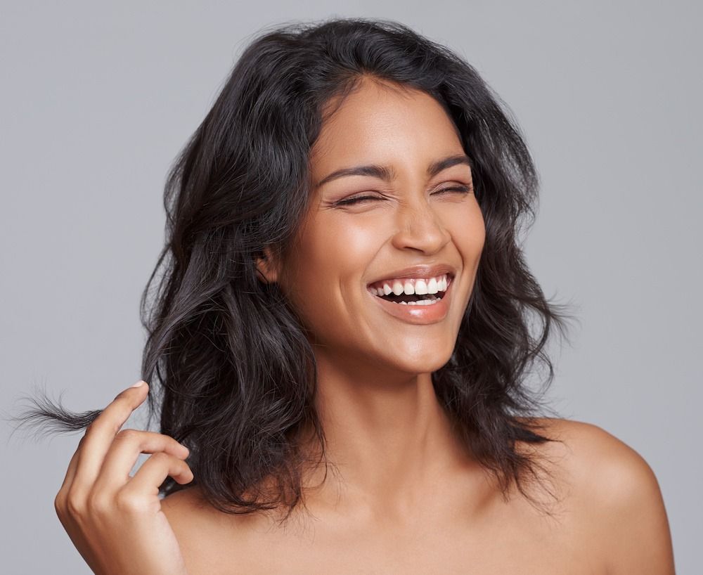 Woman with dark hair smiles, touches a strand, and looks to the side. Gray background.