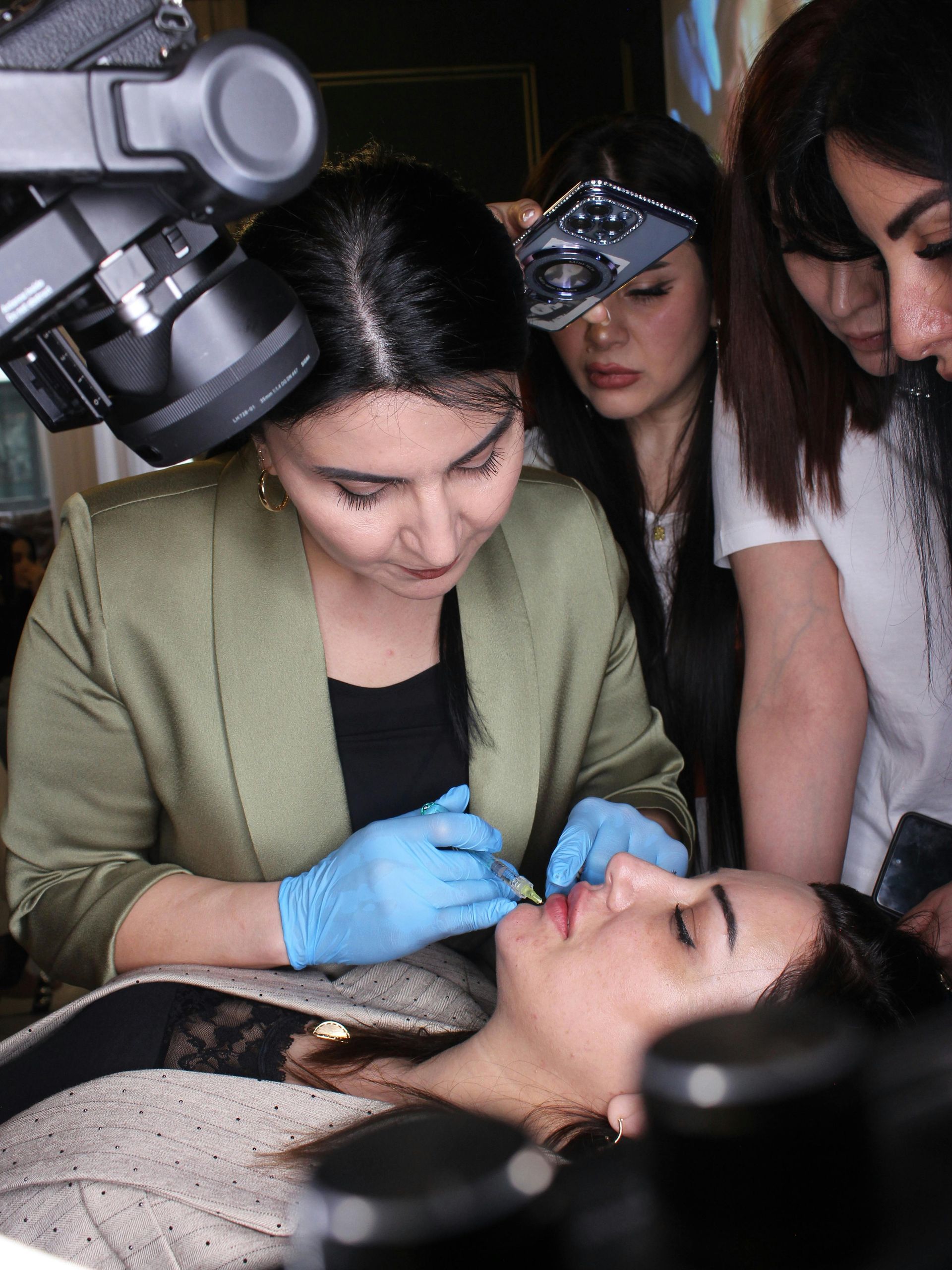 Woman in blue gloves injecting lips as students observe, lit by bright lights.