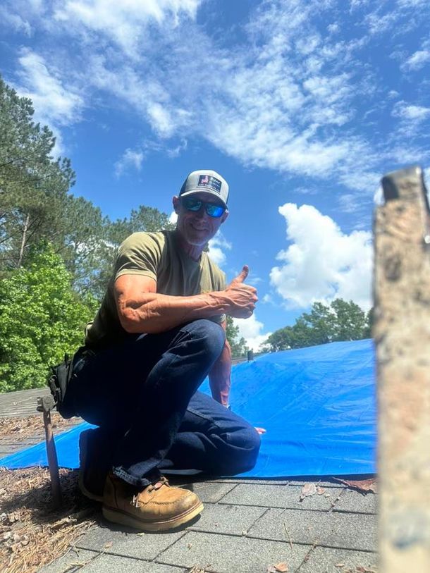 A man is kneeling on a roof with a blue tarp and giving a thumbs up.