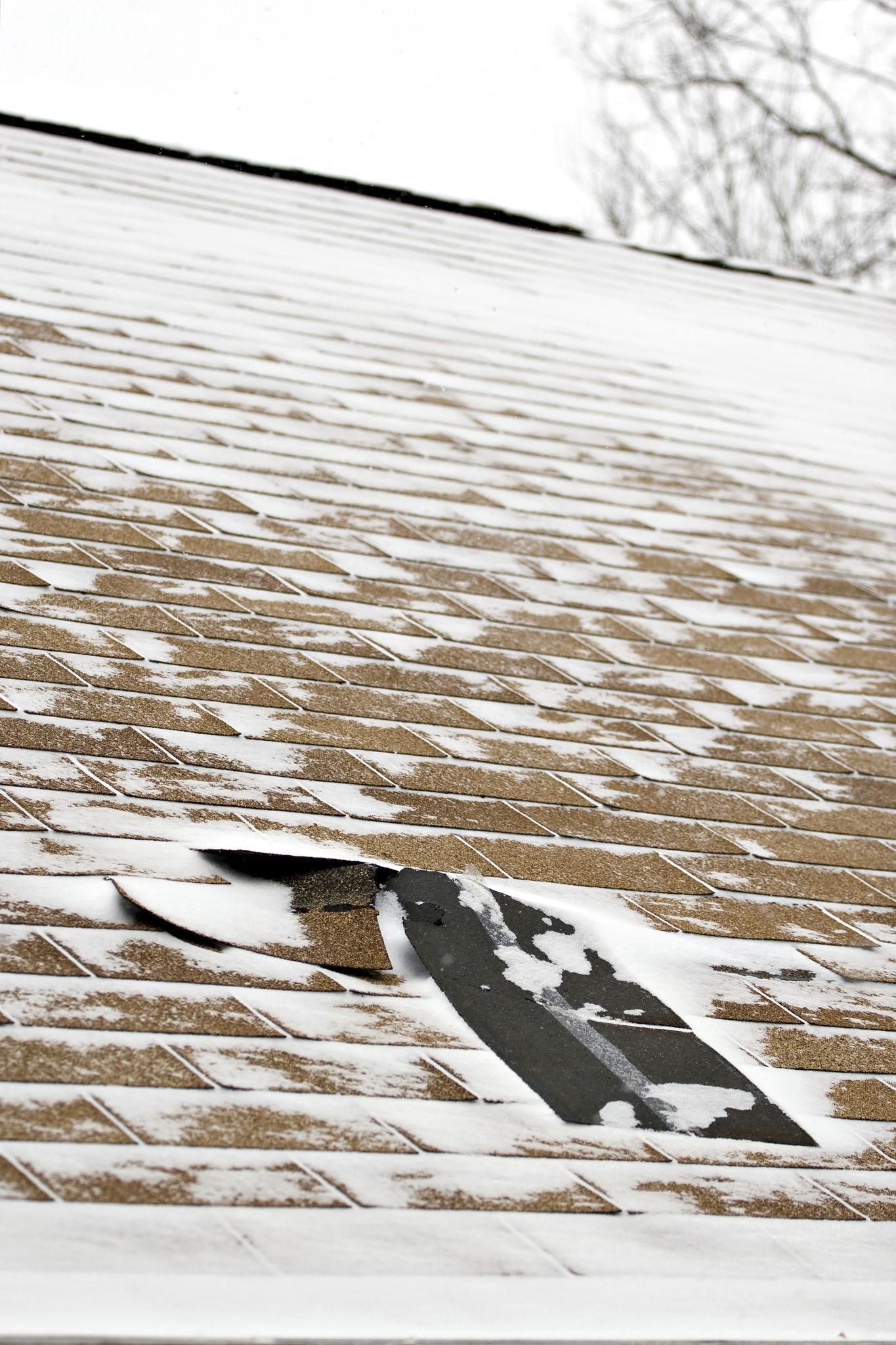 Snow-covered roof with a visible section of damaged shingles. The roof is tan, and the sky is white.