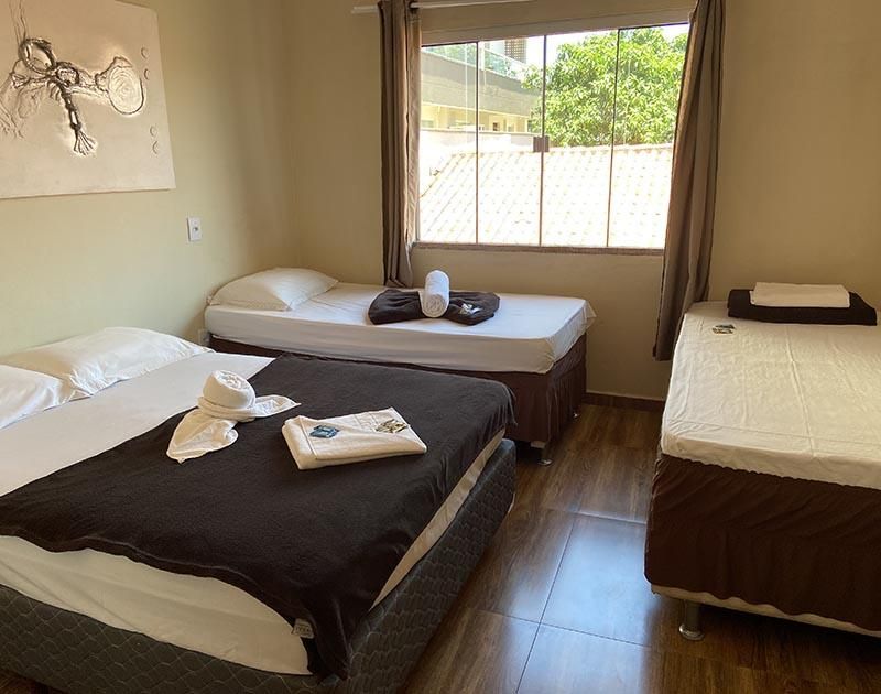 Um quarto de hotel com uma cama grande em primeiro plano, duas camas de solteiro encostadas na parede e uma janela com cortinas.