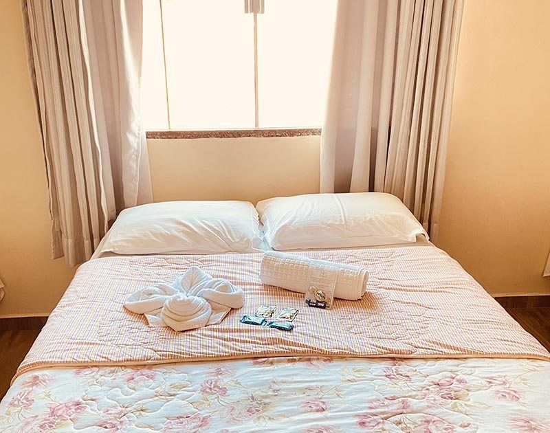 Uma cama de casal arrumada com travesseiros brancos, uma colcha floral, um arranjo de toalhas em formato de coração e artigos de higiene pessoal em um quarto iluminado.