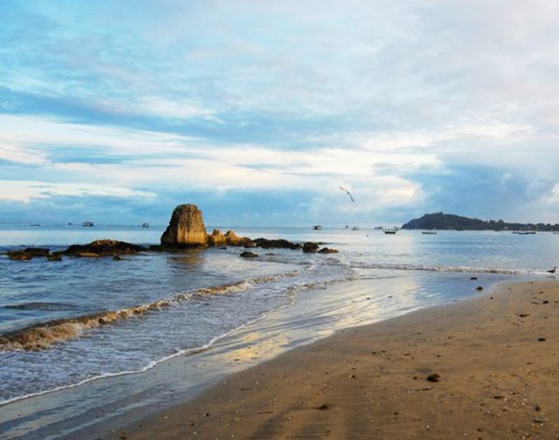 Uma praia pitoresca ao entardecer, com uma grande formação rochosa nas águas calmas sob um céu nublado azul e dourado.