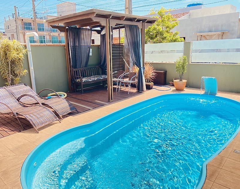 Uma piscina azul brilhante fica em um pátio ao lado de uma pérgola de madeira com assentos sombreados e duas espreguiçadeiras de vime.