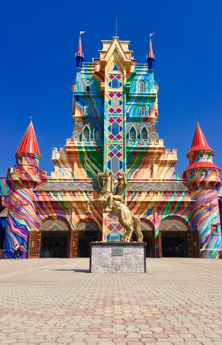 Um castelo colorido de vários andares com uma estátua equestre dourada na frente, sob um céu azul claro.