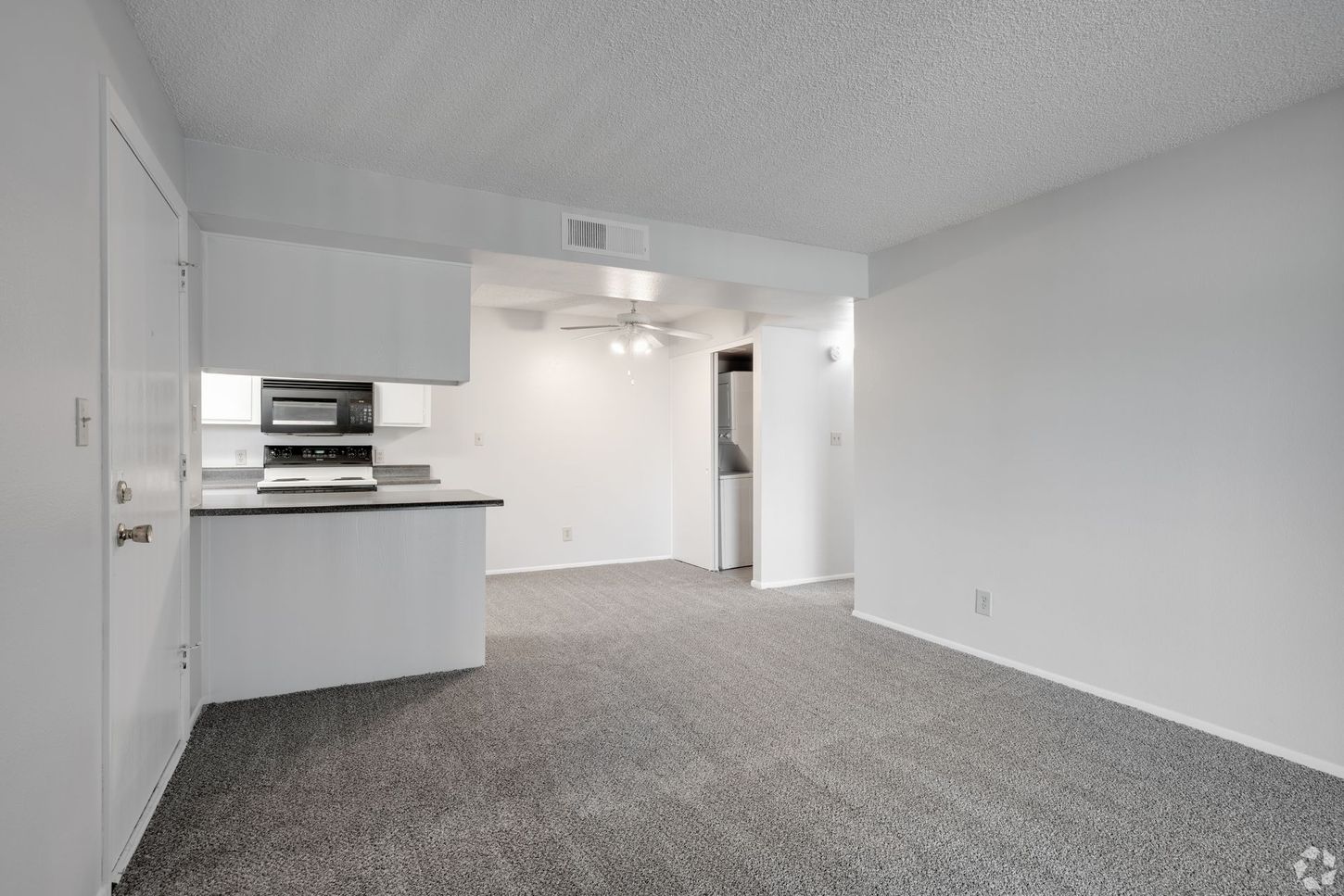 Interior view of an apartment, showing a kitchen with white cabinets, and living space with gray carpet.