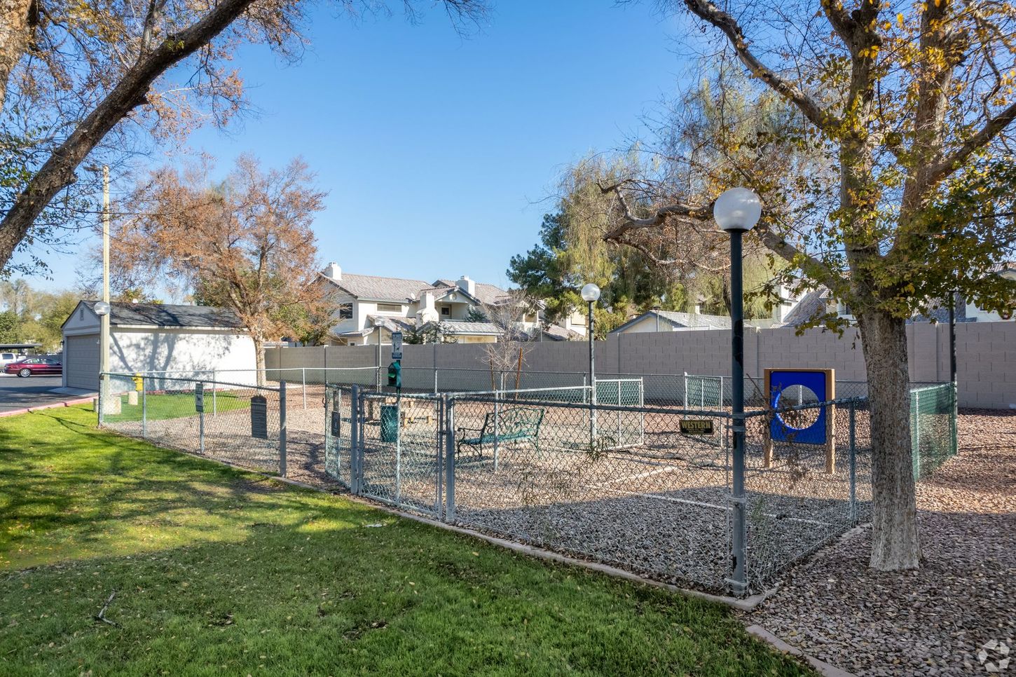 Fenced dog park with agility equipment, gravel ground, and surrounding buildings under a blue sky.