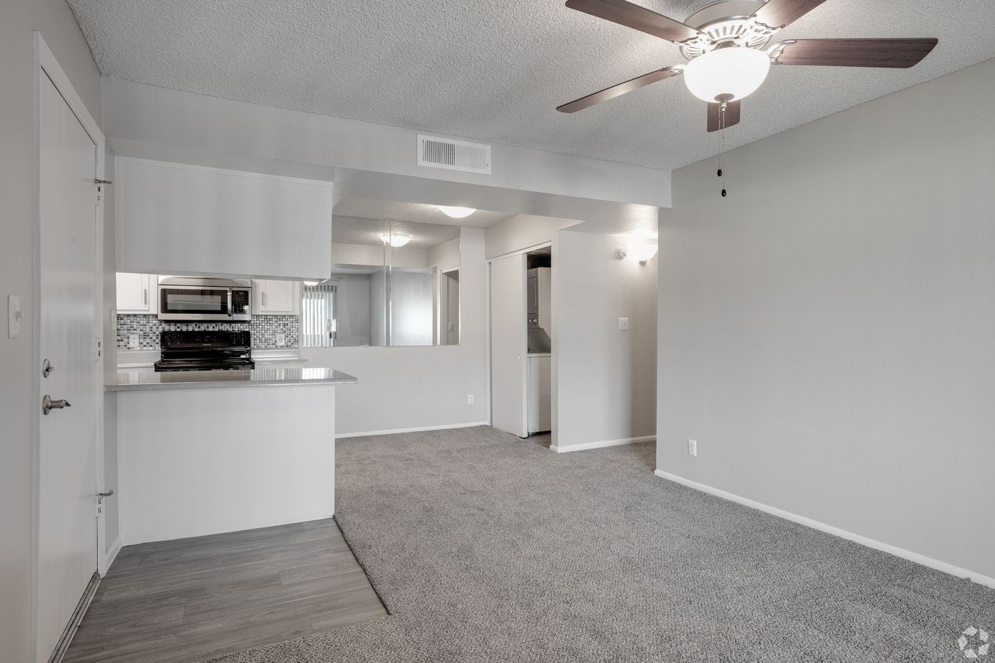 Interior of a living space with white walls, carpet, kitchen, and a ceiling fan.