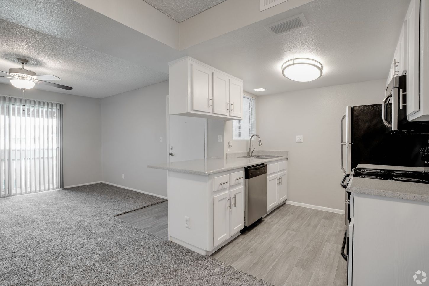Open-concept apartment interior with kitchen, living area, white cabinets, stainless steel appliances, and gray carpet.