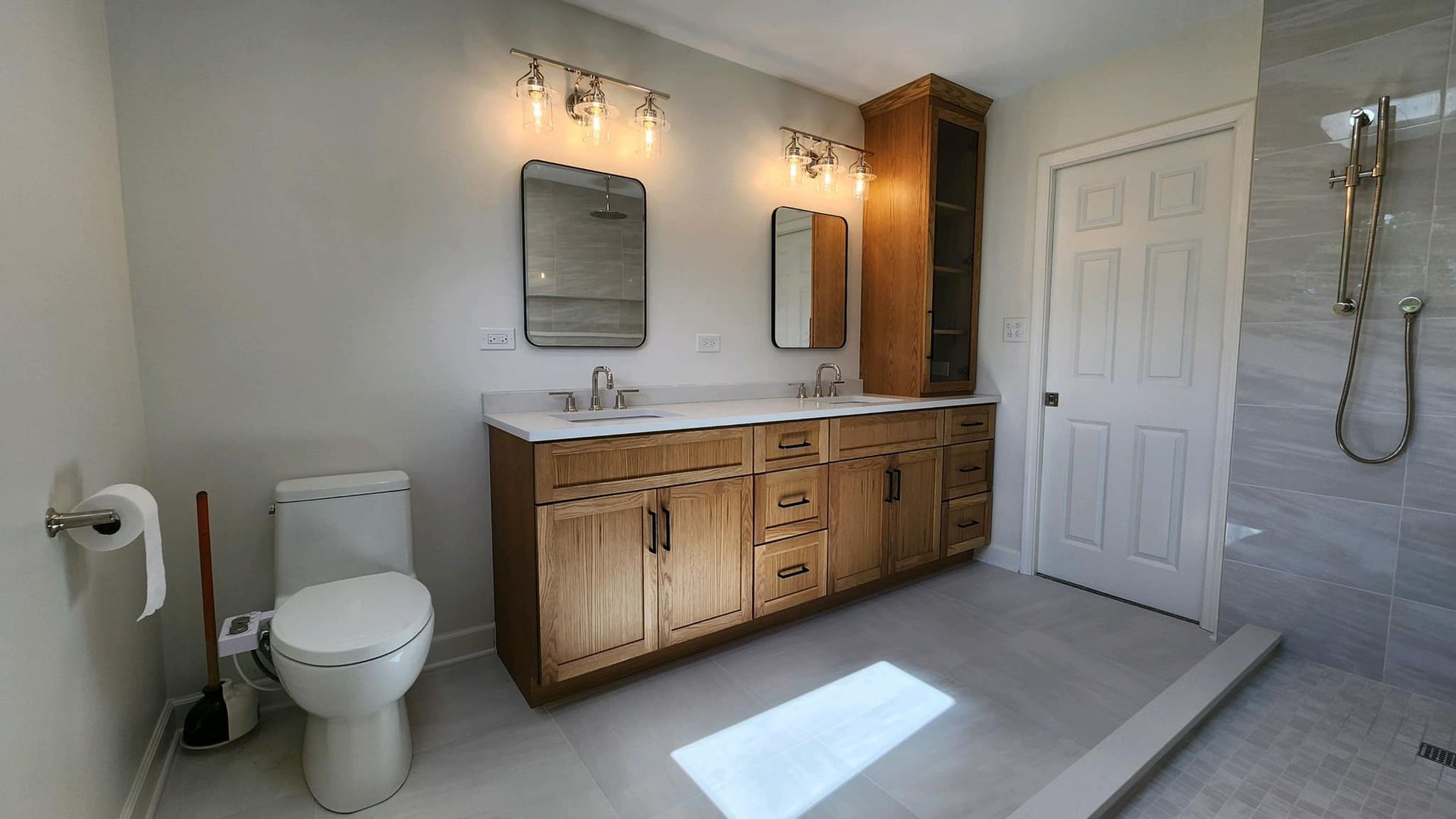 Bright bathroom with wood vanity, toilet, mirrors, shower, and sunlight on tile floor