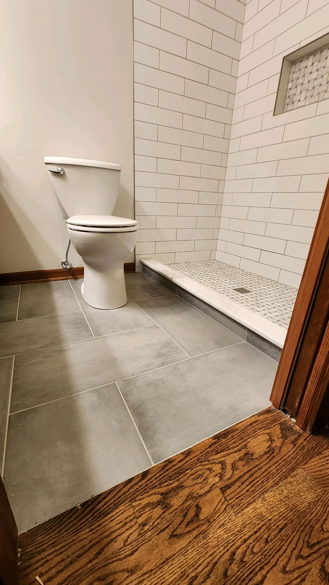 Small tiled bathroom with a white toilet, gray floor tiles, and a wooden floor transition.