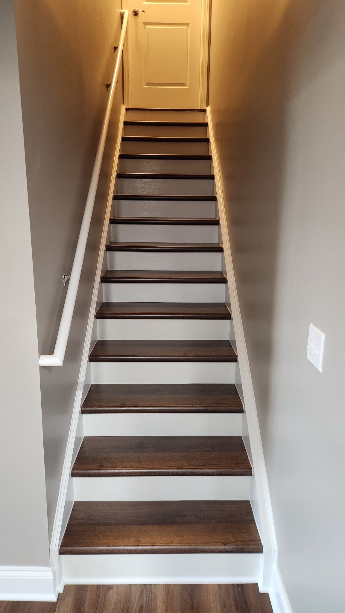 Narrow indoor staircase with dark wooden steps, white trim, beige walls, and a closed door at the top