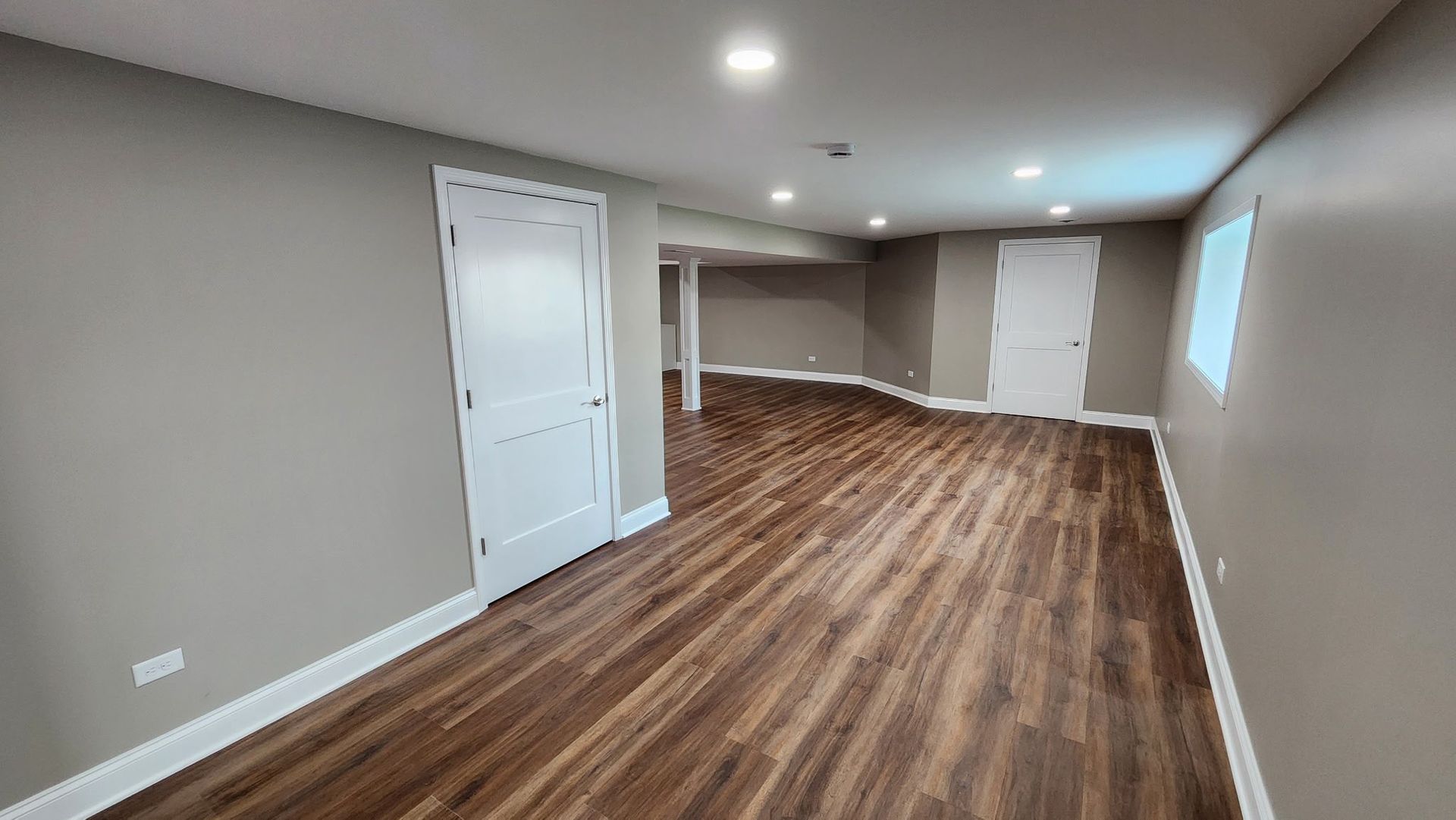 Empty finished basement with wood-look flooring, beige walls, white doors, and recessed ceiling lights.