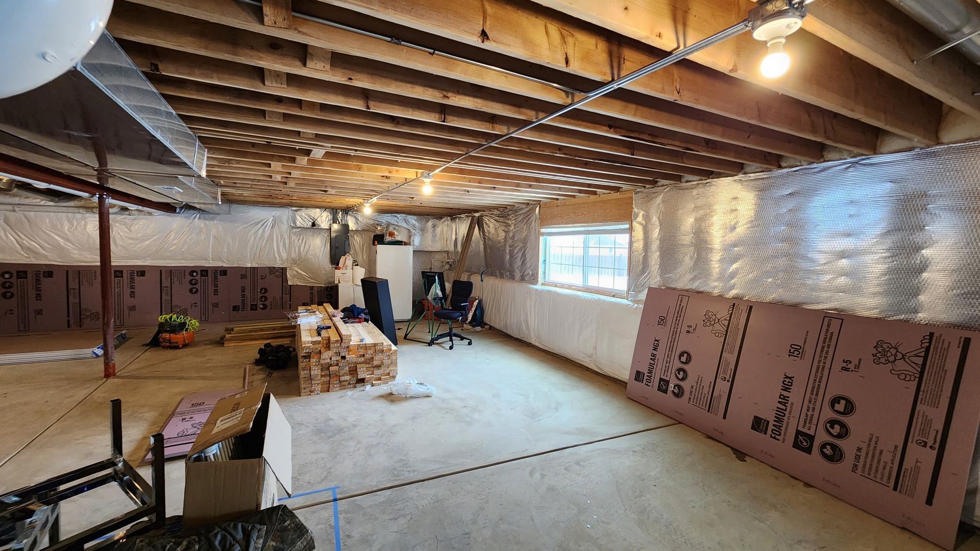 Unfinished basement with exposed beams, insulation, tools, and stacked drywall panels under warm lights