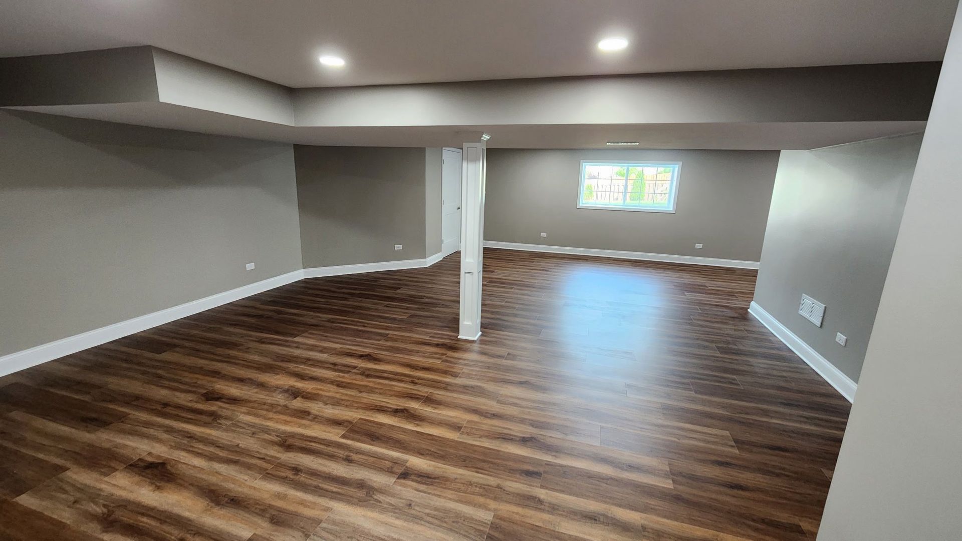 Empty finished basement with wood floors, gray walls, recessed lights, and a small window at the far end