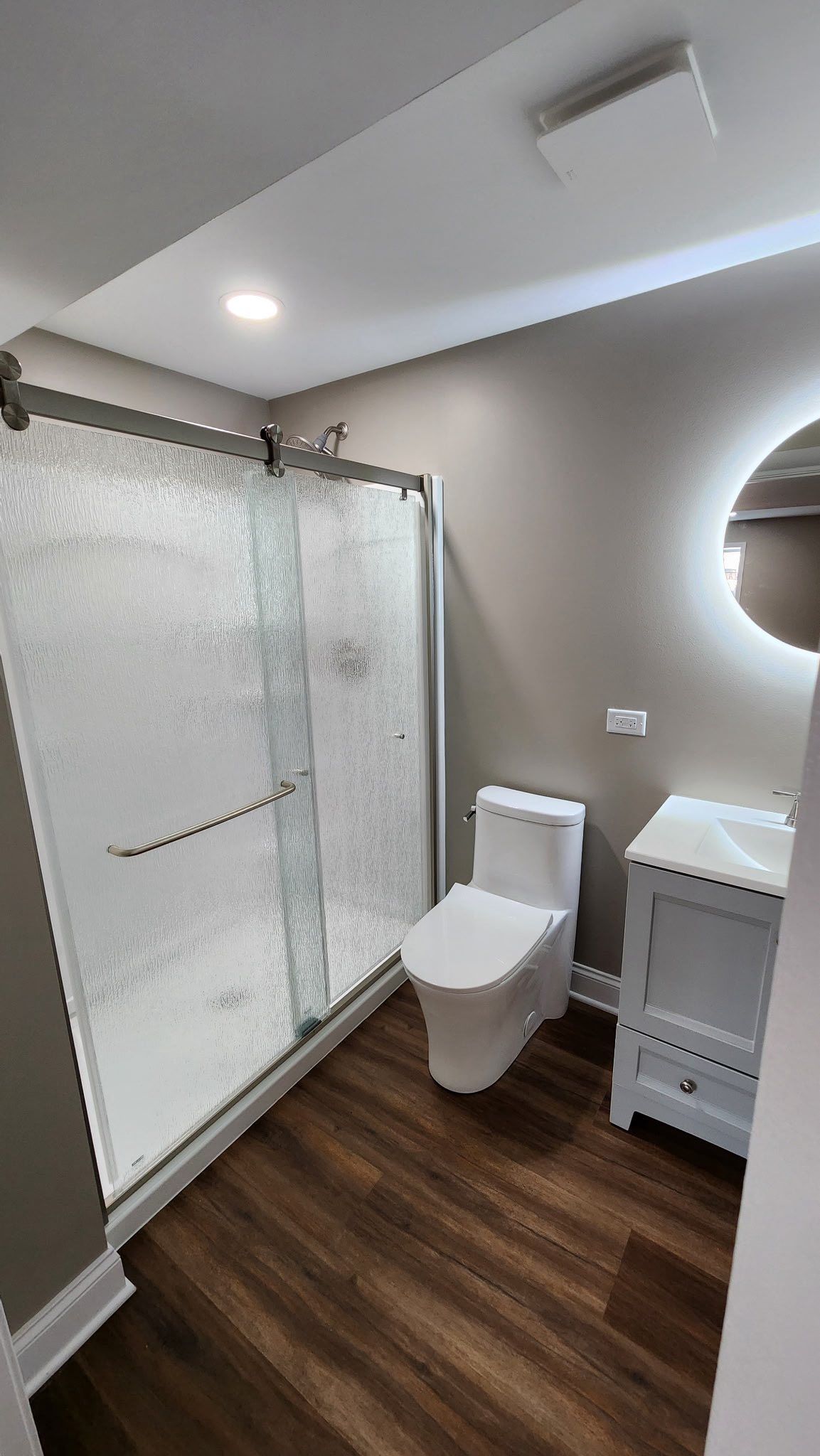 Modern bathroom with glass shower, toilet, vanity, wood-look floor, and round mirror