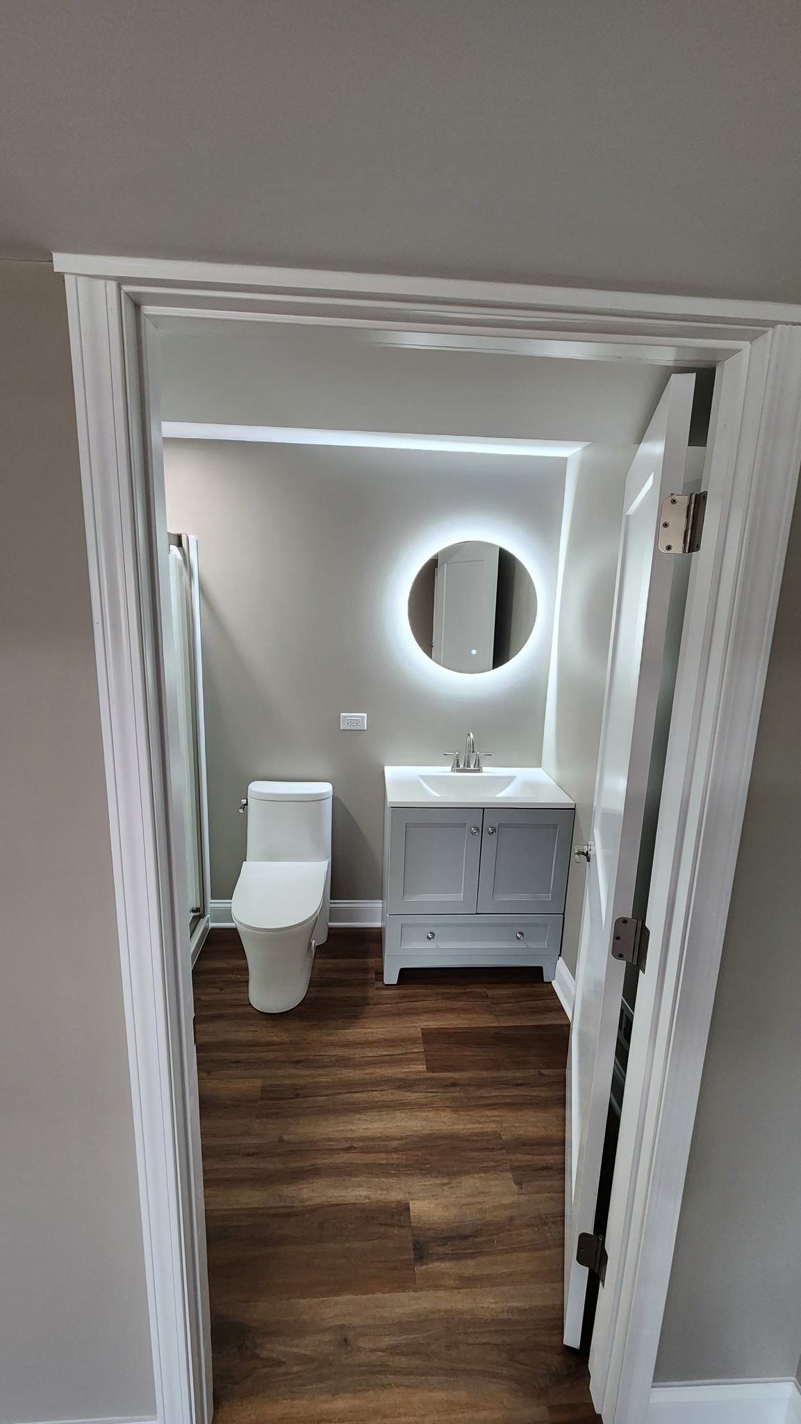 Small white bathroom with toilet, vanity, round mirror, and wood-look floor, viewed through open doorway
