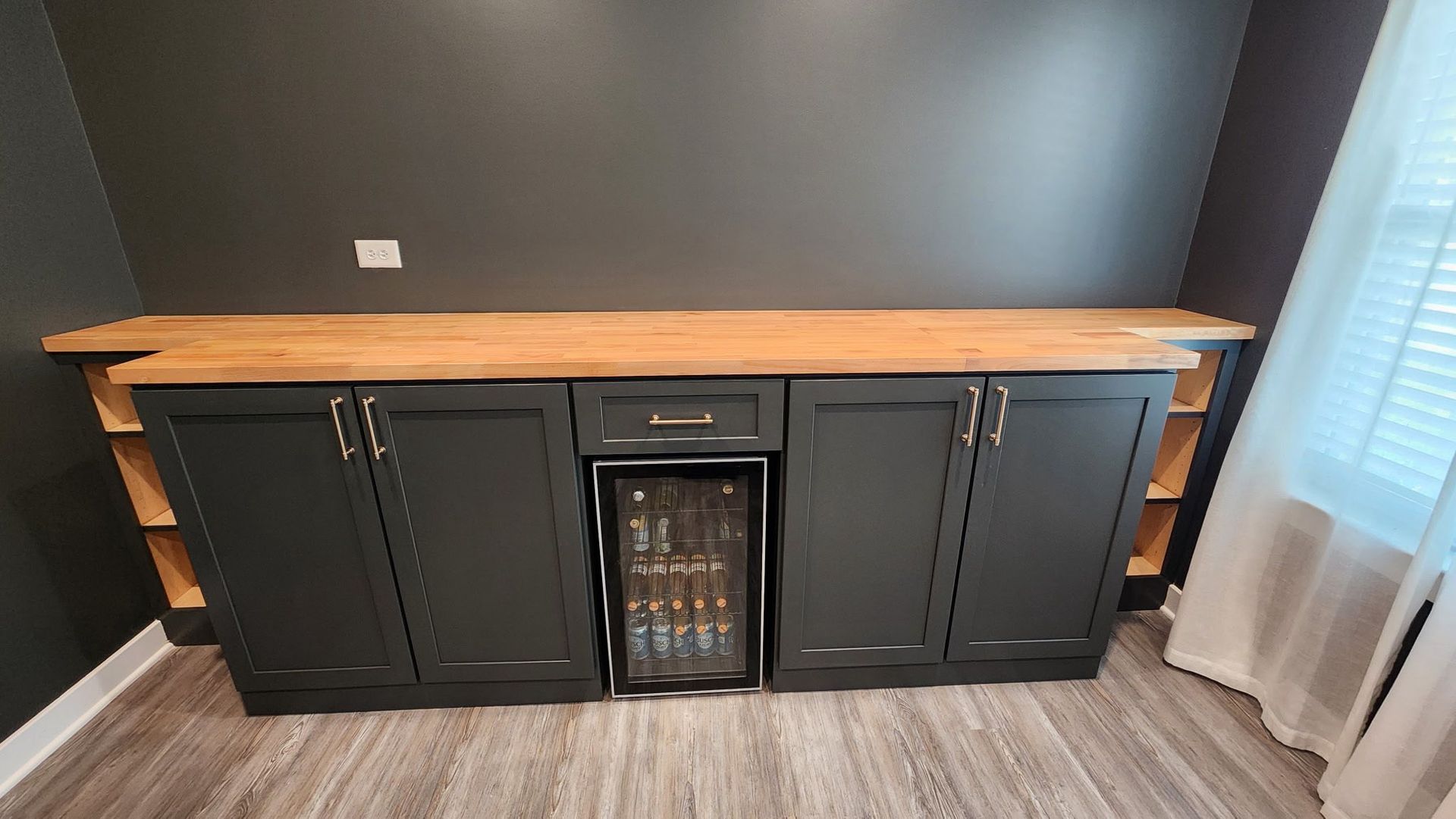 Black cabinet with wood top and wine cooler against a dark wall, beside a window with sheer curtains.