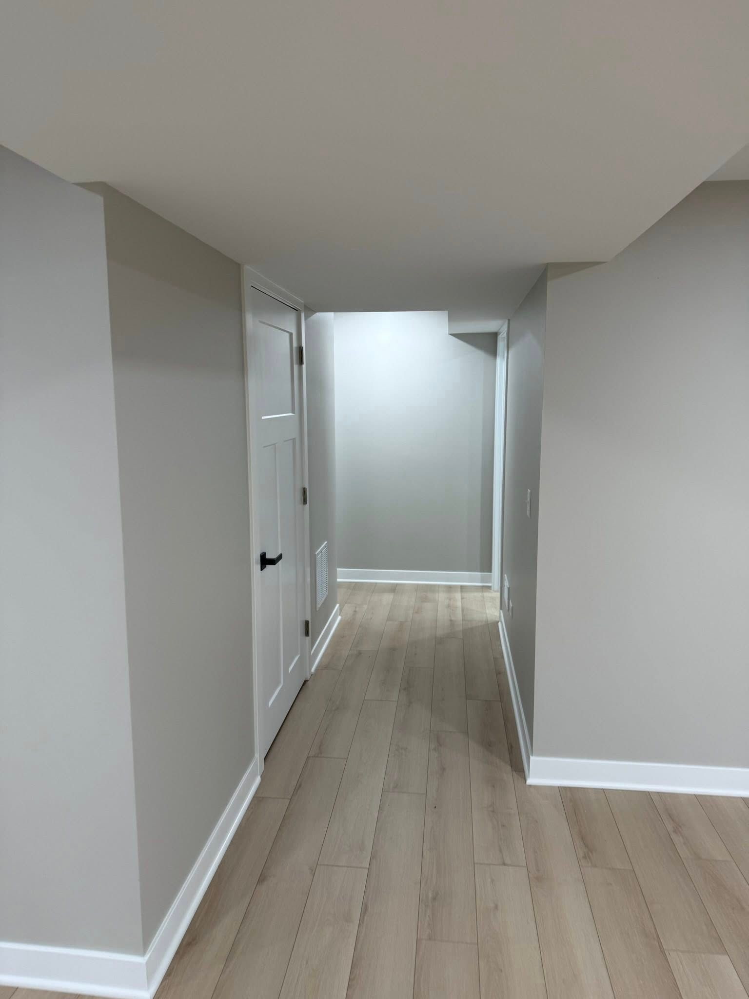 Narrow hallway with gray walls, white doors, and light wood flooring leading to a brighter room