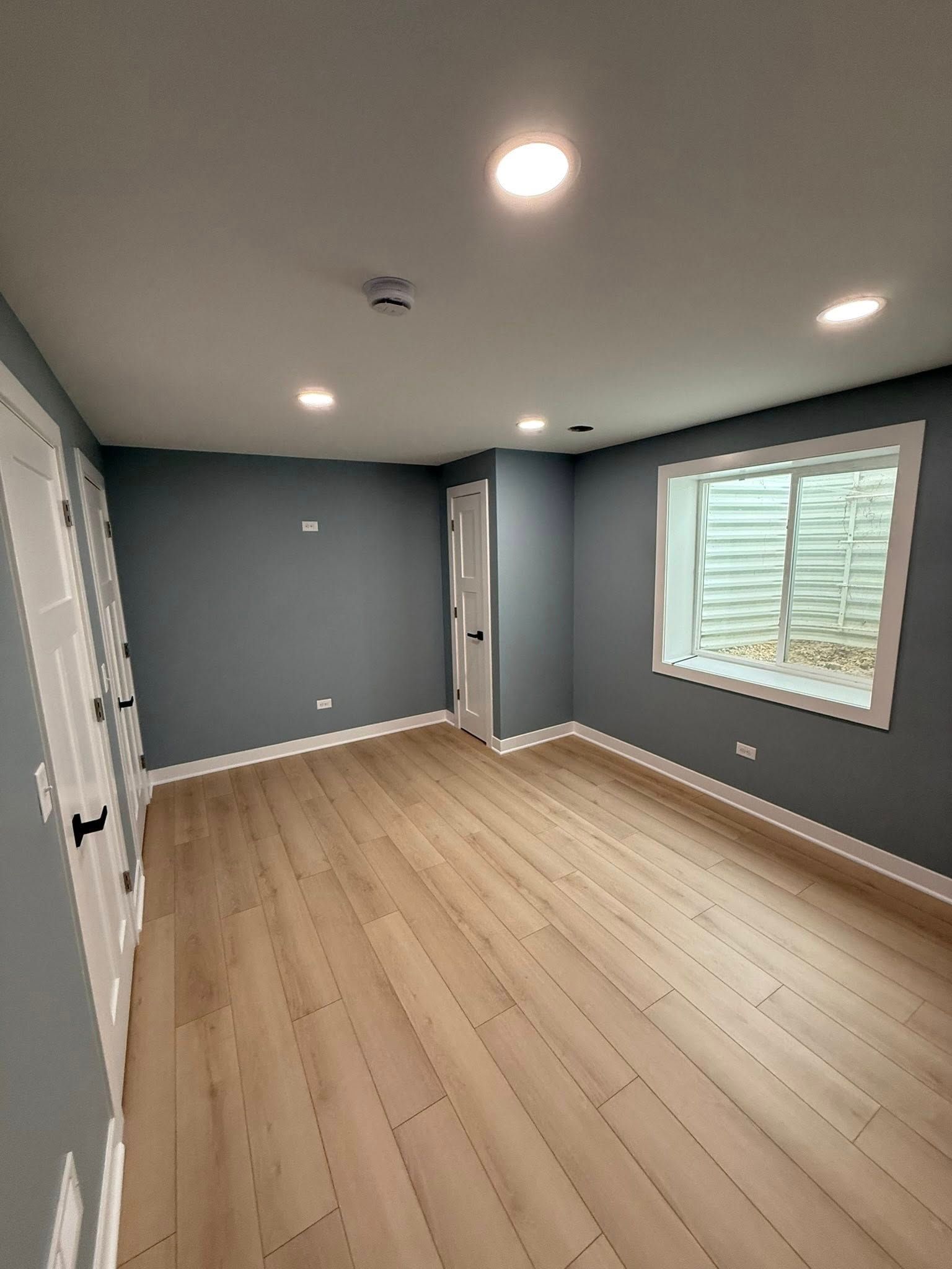 Empty gray room with light wood floors, recessed lights, and a window with blinds