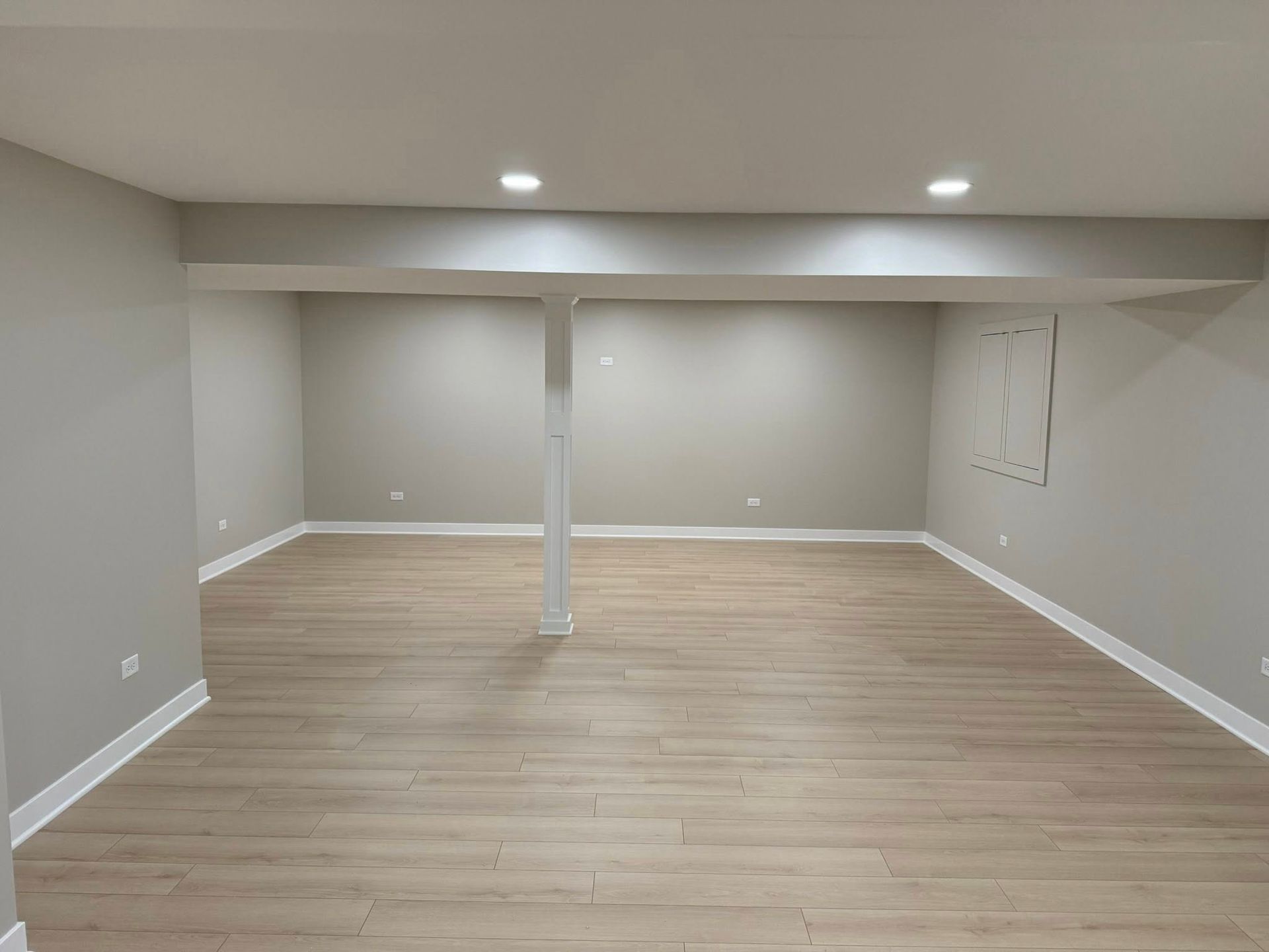 Empty finished basement room with beige walls, light wood floor, recessed lights, and a support column.