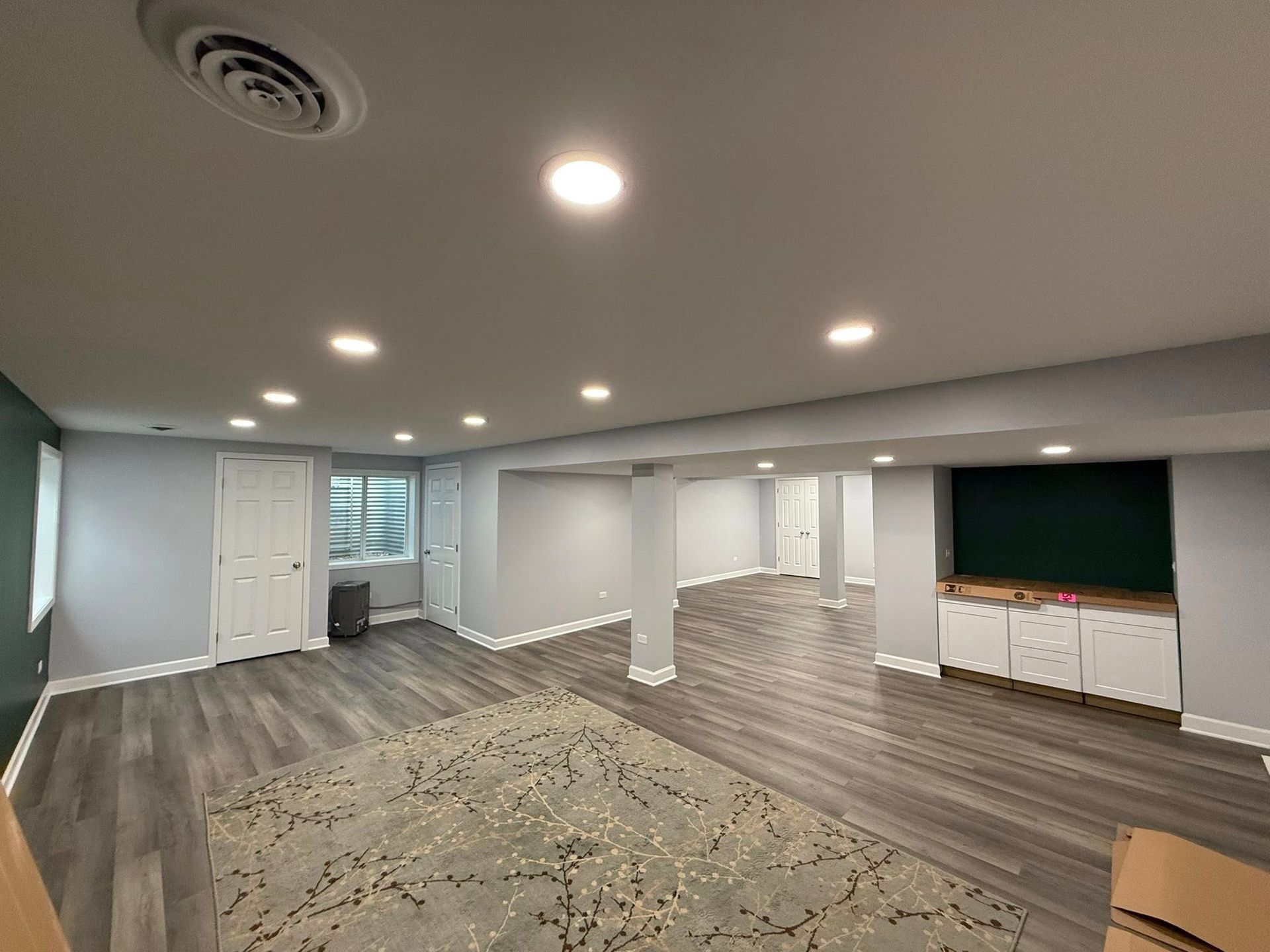 Empty basement room with gray walls, wood floors, recessed lights, and a large rug, viewed from the corner