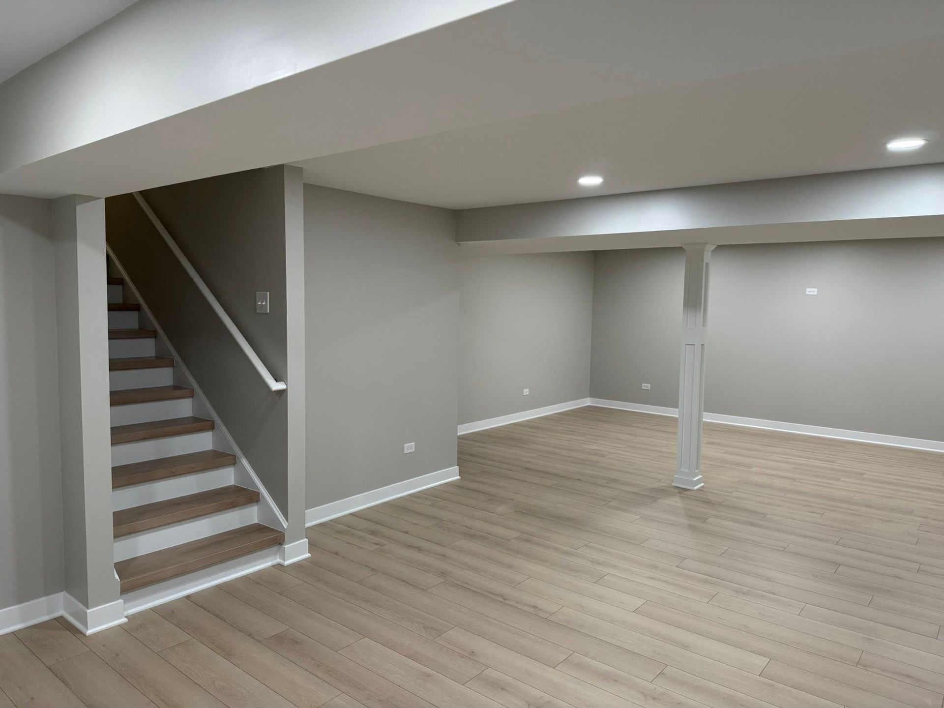 Empty finished basement with gray walls, light wood floors, stairs, and a support column.