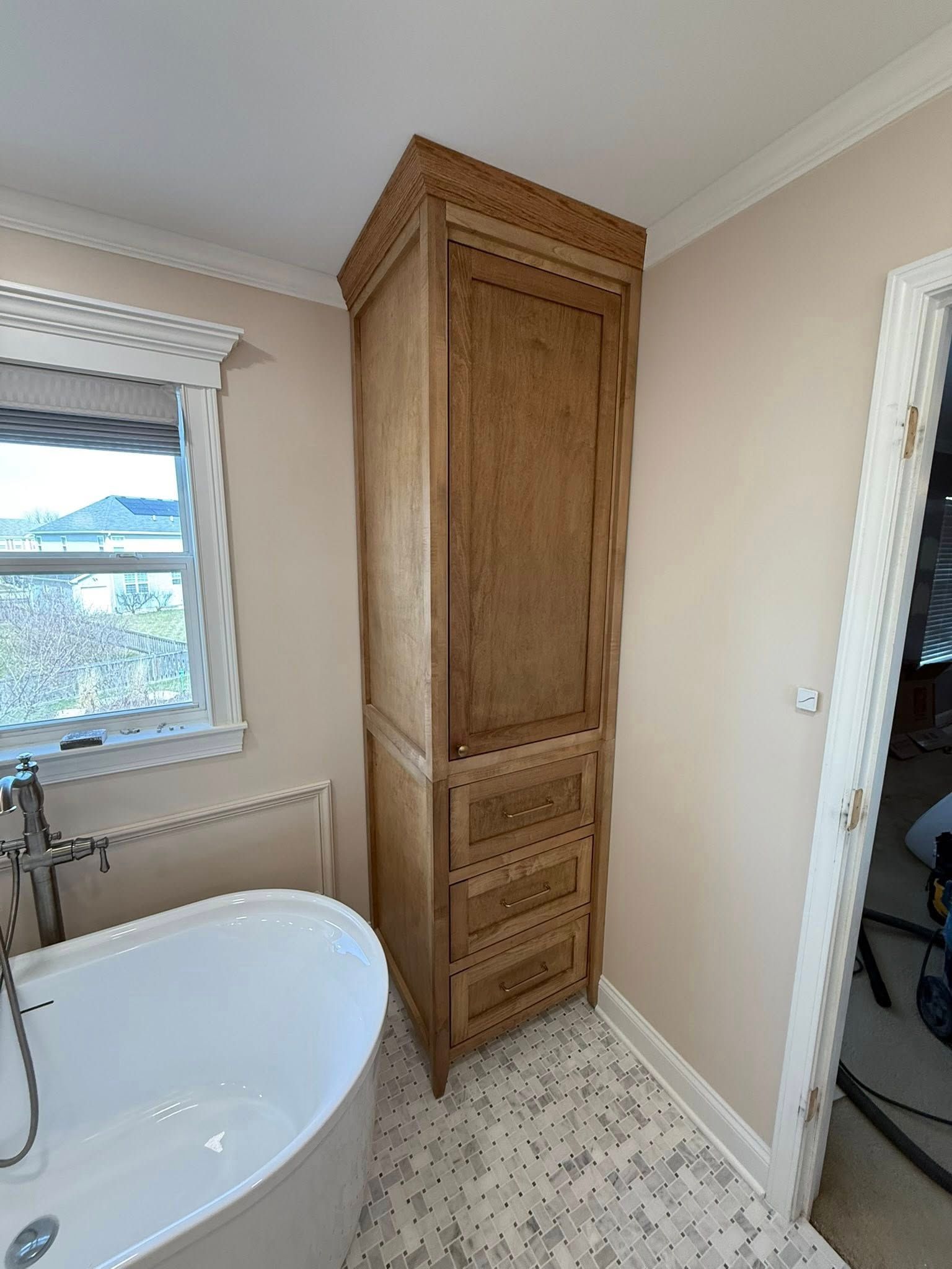 Tall wooden cabinet in a beige bathroom beside a bathtub and window.