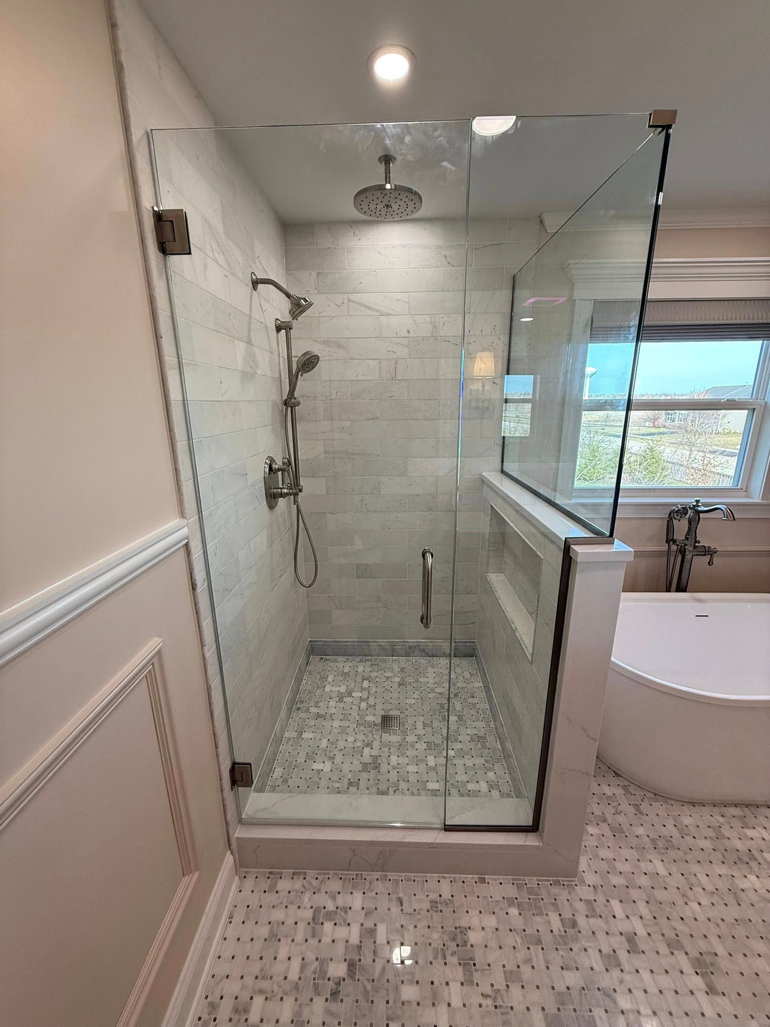 Spacious tiled bathroom with glass walk-in shower, white tub, and window light.