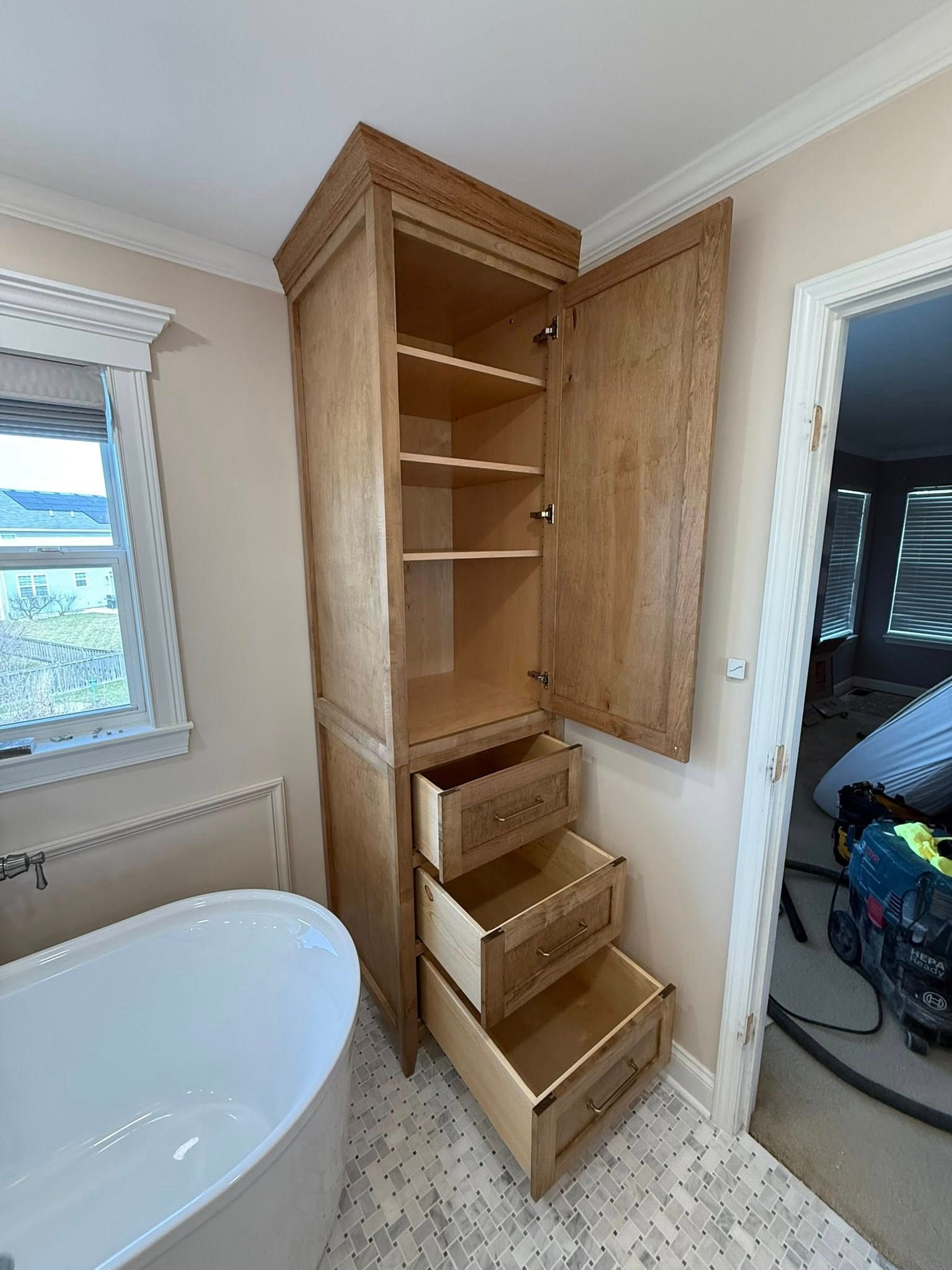 Tall wooden bathroom cabinet with shelves and drawers beside a bathtub and doorway.