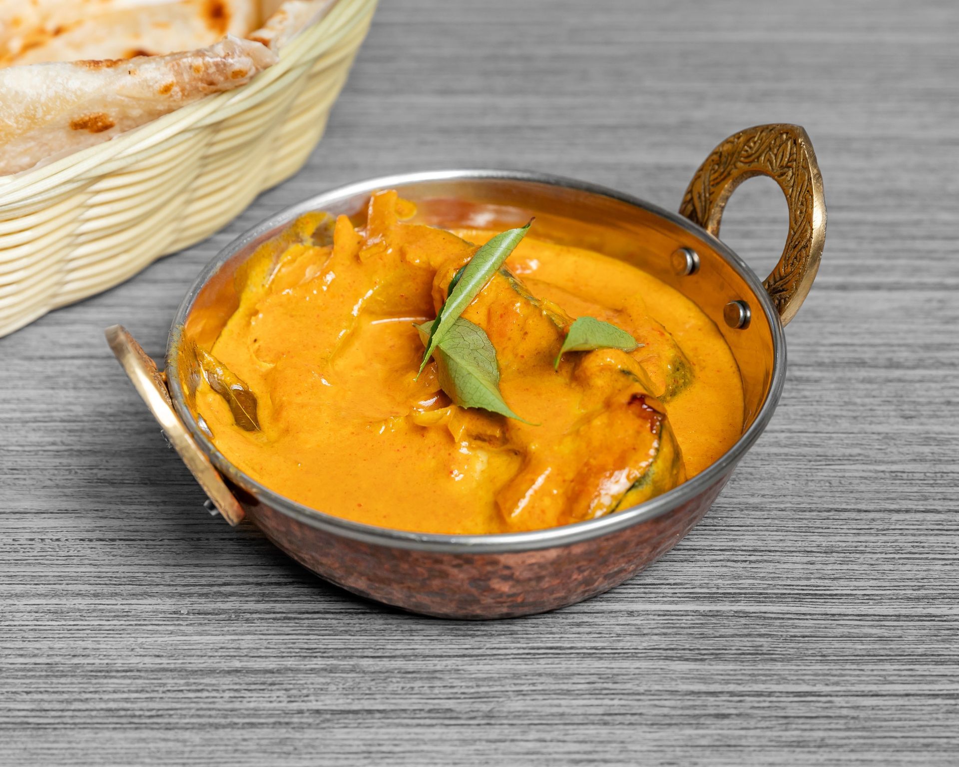 A bowl of fish curry is on a table next to a basket of roti