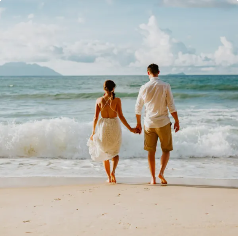 A man and a woman are walking on the beach holding hands.