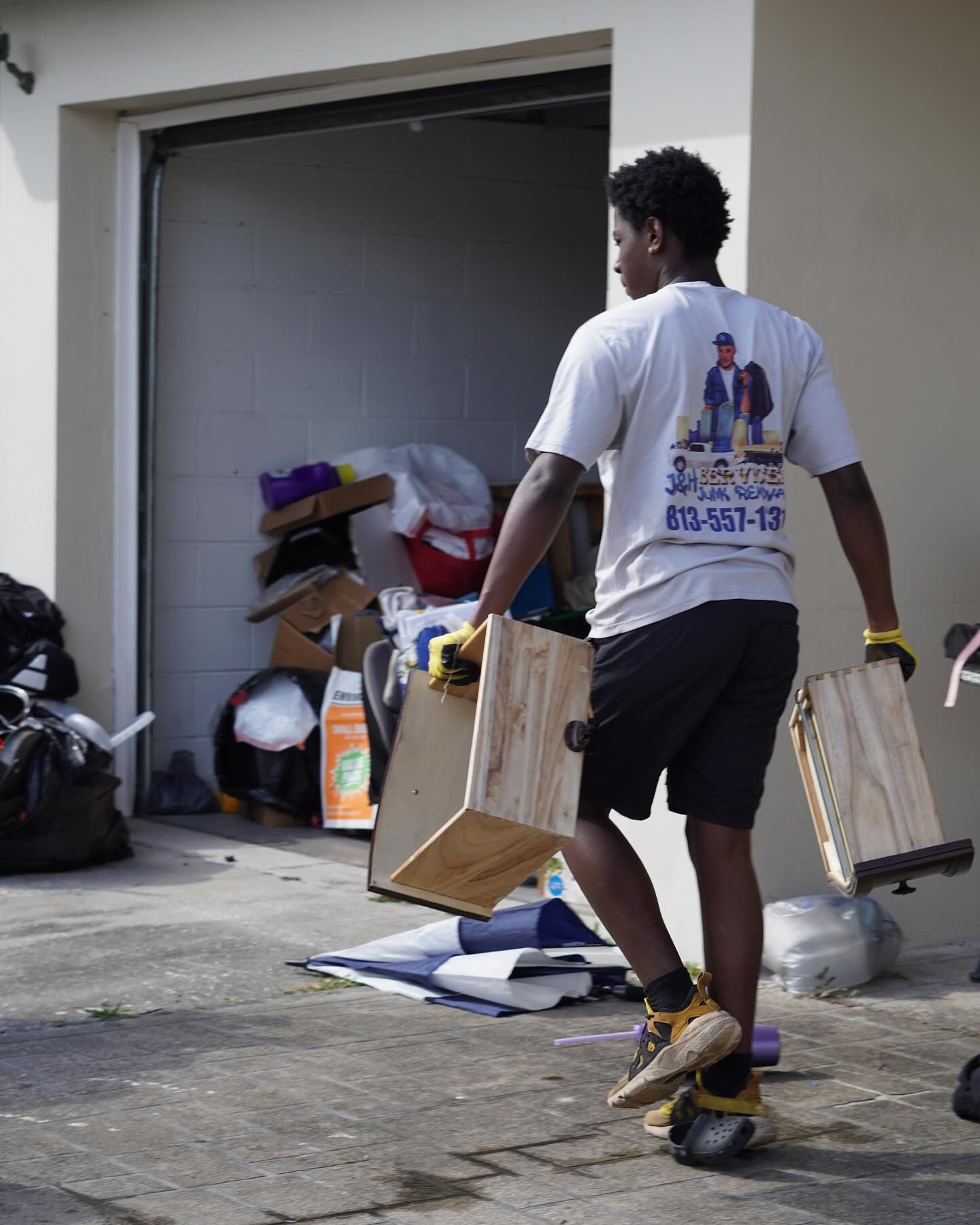 Man in white t-shirt carries wooden boxes out of a garage. Boxes, trash bags, and boxes are behind him.