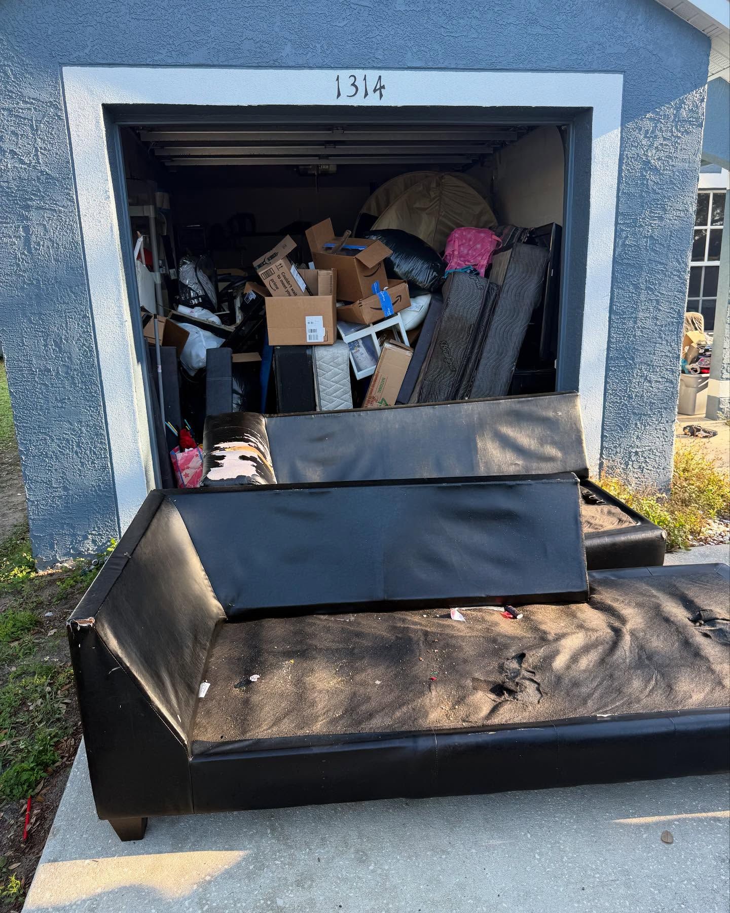 A cluttered garage with a damaged black couch in front. Boxes and various items fill the space.
