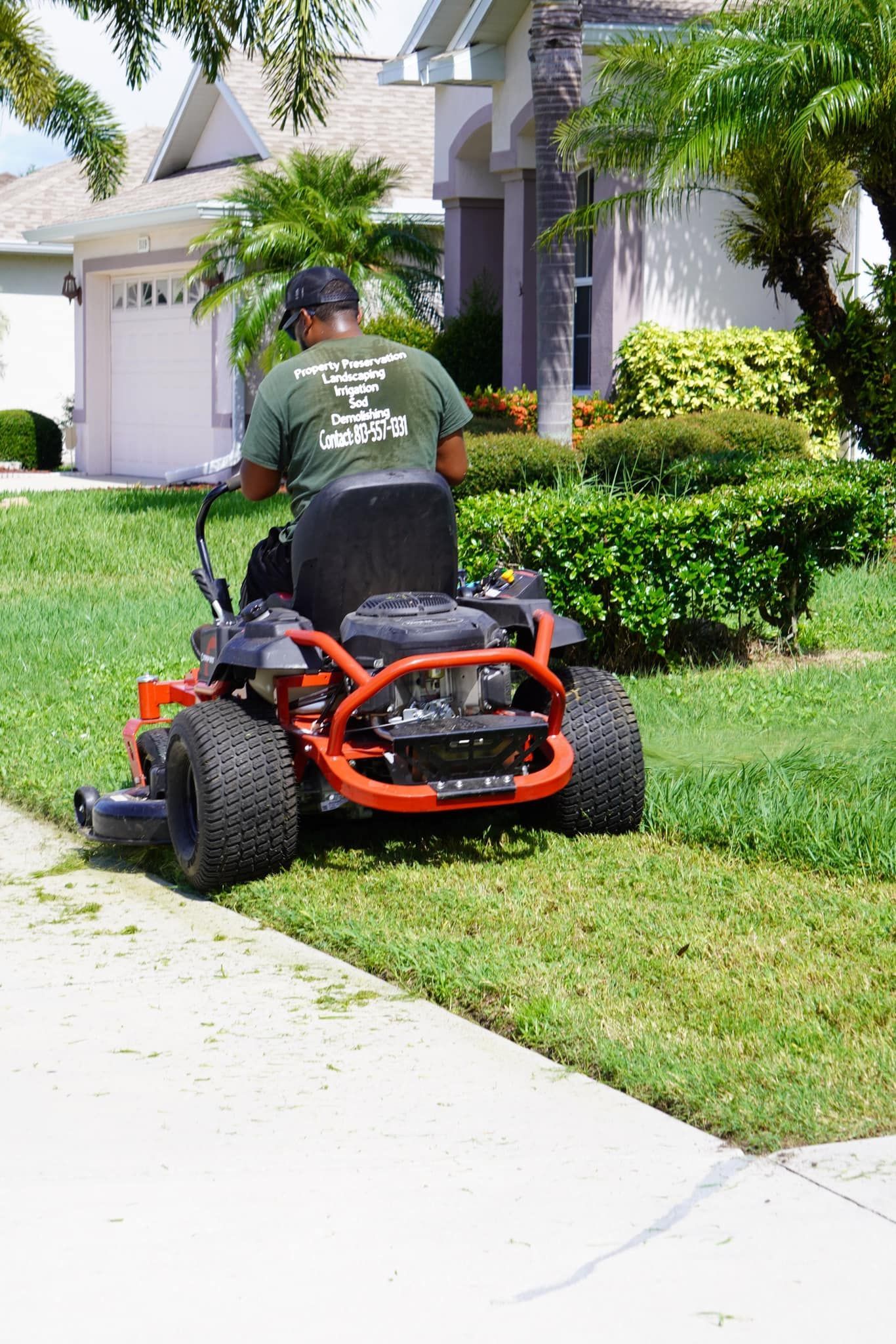 A man on a red zero-turn mower mows grass along a sidewalk in front of a house.