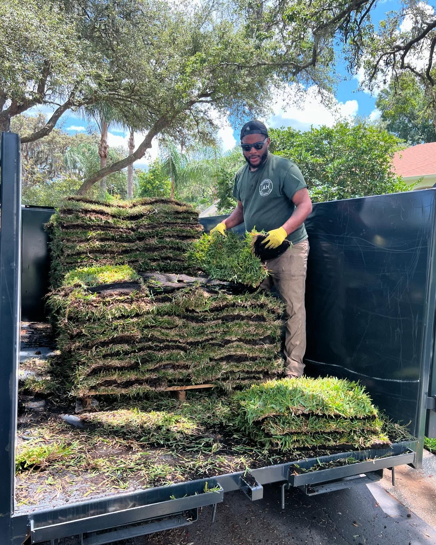 Man in green shirt, khaki pants, and gloves, stacks sod in a truck bed on a sunny day.