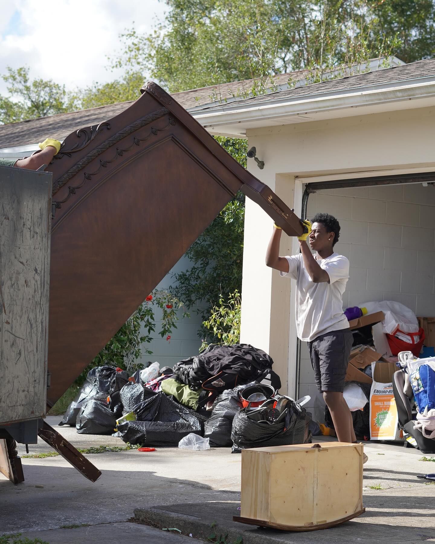 Man lifting debris into a dumpster in front of a garage. Black bags and a wooden box are in front.