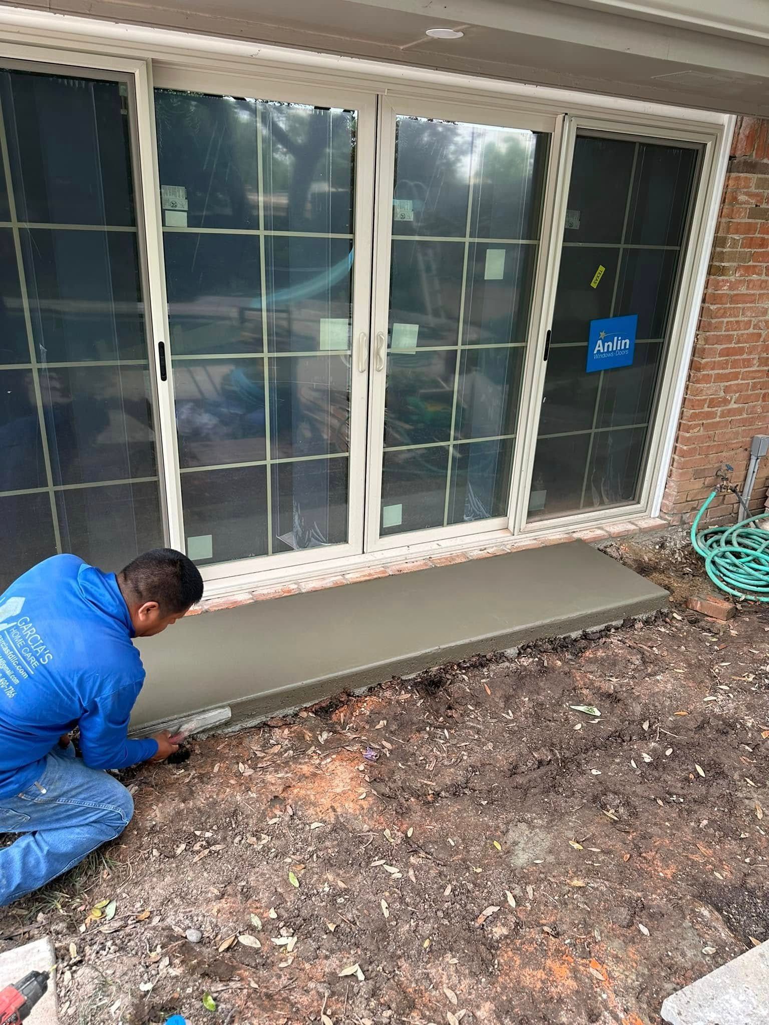 A man is kneeling on the ground working on a concrete walkway in front of a sliding glass door.