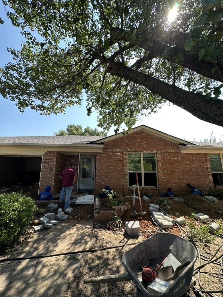 A man is standing in front of a brick house with a wheelbarrow in front of it.