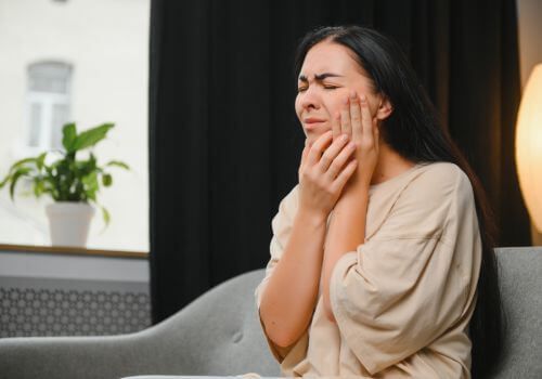 Person holding face with eyes closed, sitting on a couch in a dim room with a plant and lamp nearby.