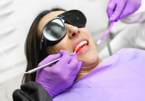 Dentist examining a patient’s mouth with tools; patient wears sunglasses and purple bib.