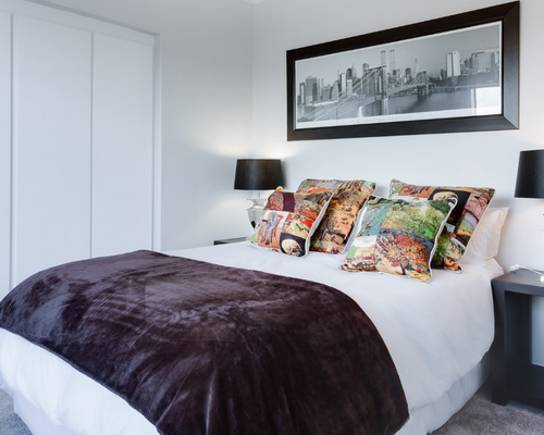 Modern bedroom with white walls, bed with dark throw, colorful pillows, and framed cityscape art above the headboard